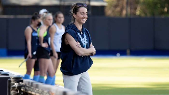 UNC field hockey coach Erin Matson smiles while watching practice with players standing behind her.